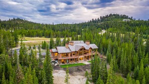 Un grand chalet en bois, entouré d’une forêt dense, avec une imposante cheminée en pierre et de nombreuses fenêtres.