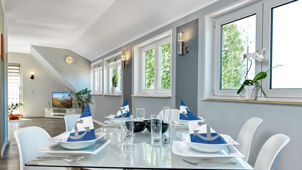 A modern dining area with a glass table set for six, white chairs, and a view of the outdoors through large windows. - Apt. WEITER BLICK - Welcome, make yourself comfortable! (Insel Poel / OT Neuhof)