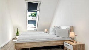 A modern bedroom with a sloped ceiling, a wooden bed frame, white bedding, a nightstand with a lamp, and a potted plant. - Apt. WEITER BLICK - Welcome, make yourself comfortable! (Insel Poel / OT Neuhof)