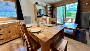 A wooden dining table set with plates, glasses, and cutlery, surrounded by chairs. A television and kitchen area are visible in the background. - T2 Apartment with Large Terrace and Pets Allowed (Samoëns)
