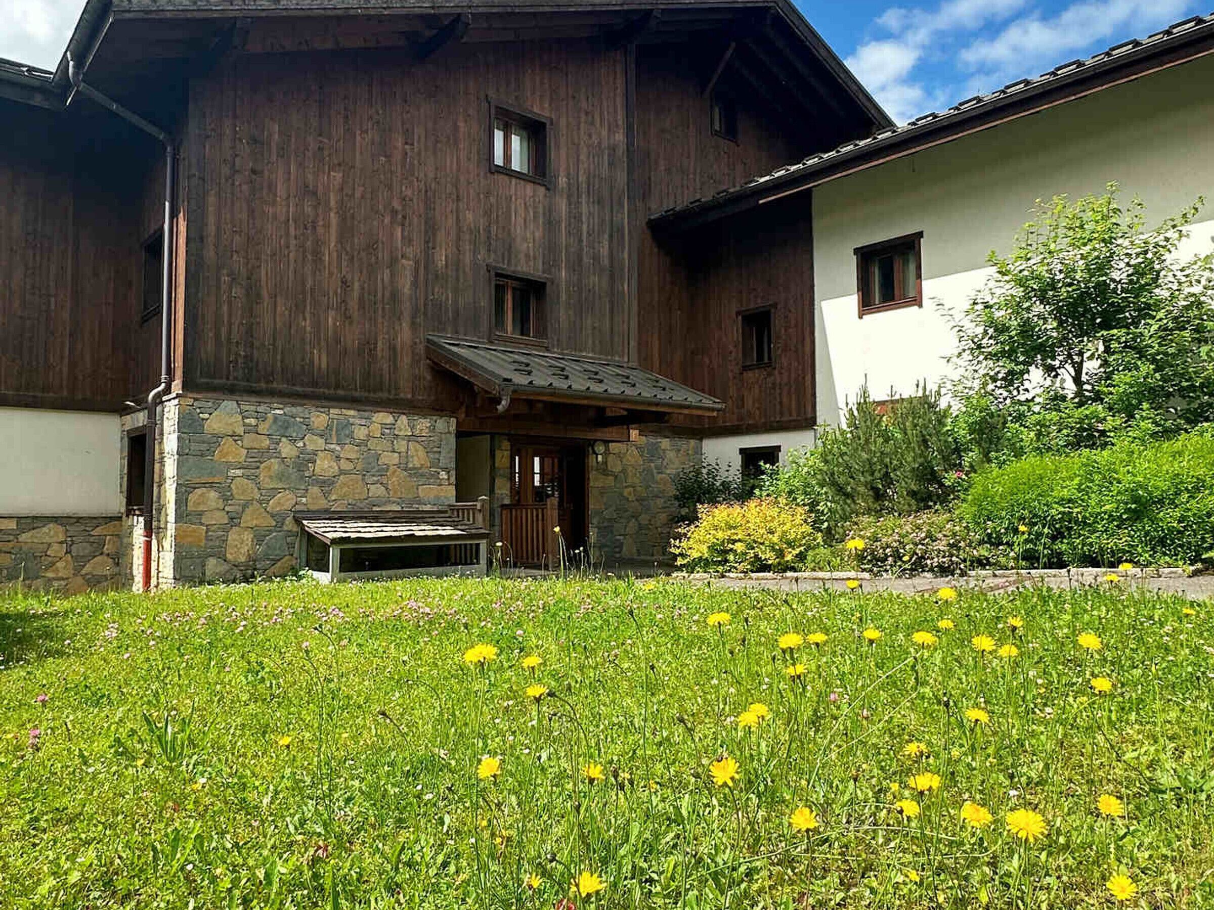 A wooden house with a stone foundation, a porch, and a grassy yard with yellow flowers.