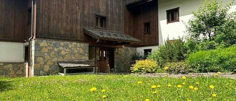 A wooden house with a stone foundation, a porch, and a grassy yard with yellow flowers.