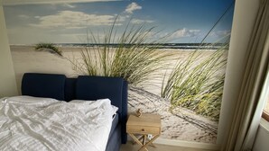 Een slaapkamer met een bed, een klein houten nachtkastje en een strandschildering op de muur.