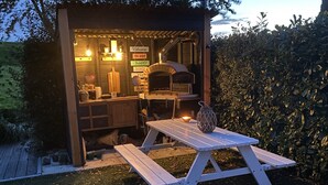 Een houten buitenkeuken met een pizzaoven, een tafel en banken.