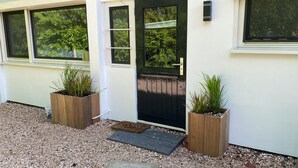 Een huisentree met een zwarte deur, twee houten plantenbakken met planten, een deurmat en een grindpad.