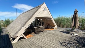 A triangular wooden structure with a thatched roof, a table with chairs, and a bicycle. - Reed hut / pile dwelling on Lake Neusiedl (Ruster Bucht)