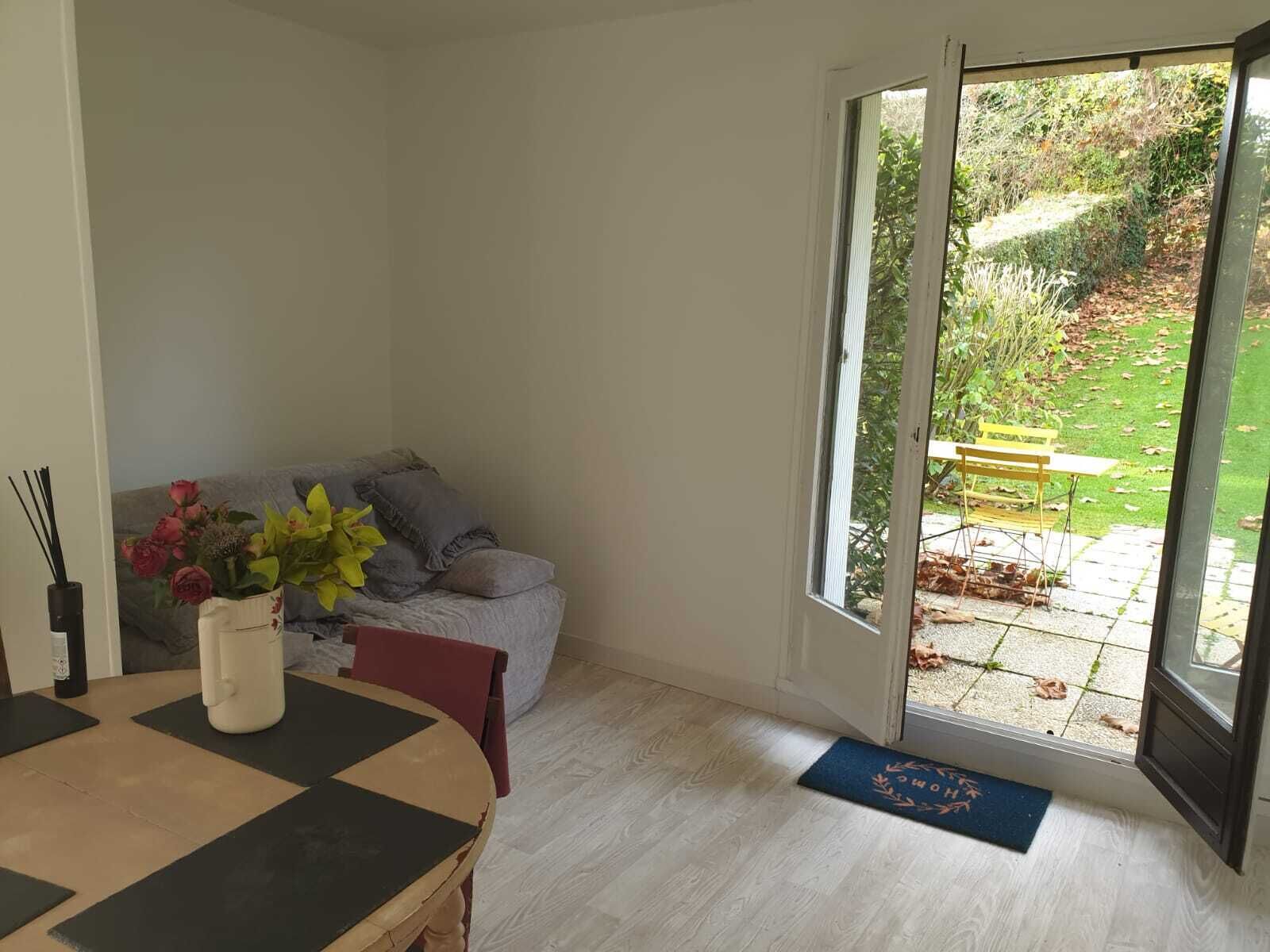 A room with a dining table, a sofa, and a view of a garden through a sliding glass door.