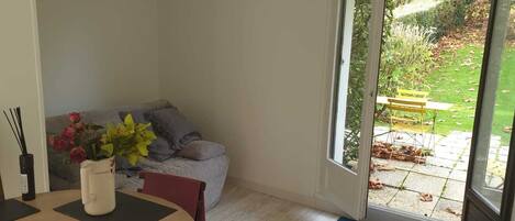 A room with a dining table, a sofa, and a view of a garden through a sliding glass door.