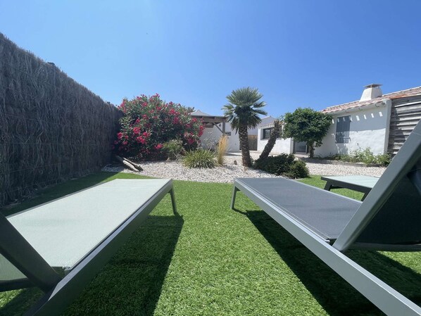 Un jardin avec une pelouse artificielle, deux chaises longues, un palmier et une maison à l’extérieur blanc.