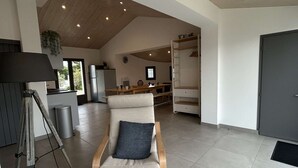 A spacious living area with a high ceiling, a wooden ceiling, a sofa with a cushion, a dining table, a kitchen area, and a wardrobe.