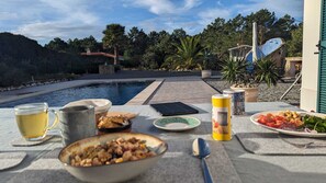 Taula de menjador amb menjar, begudes i una tauleta digital a la piscina, amb vista a arbres i un edifici.