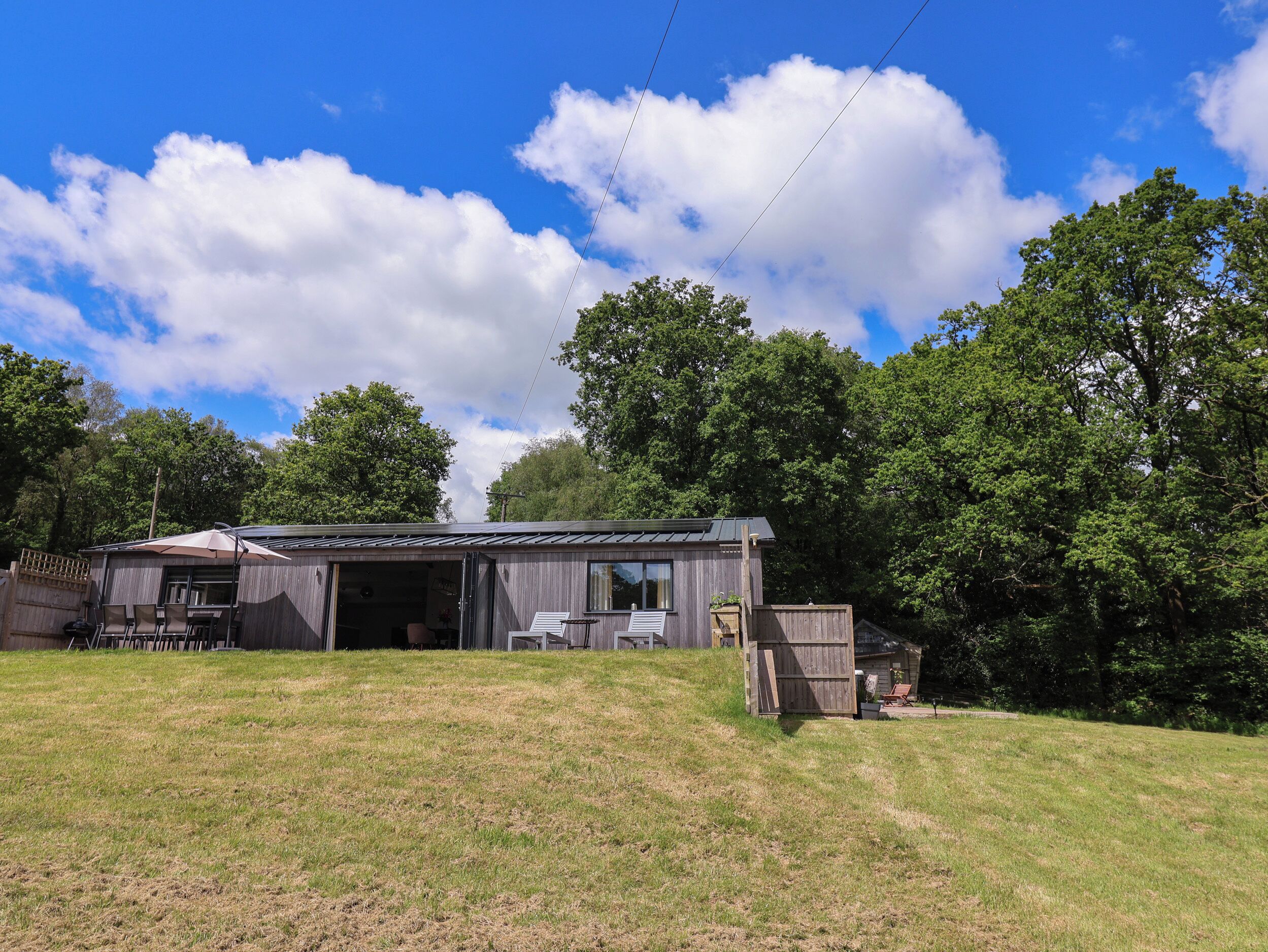A rustic cabin with a large open garage, surrounded by a grassy yard and dense trees.
