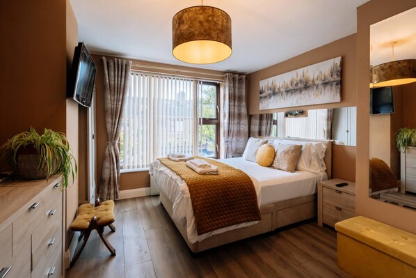A modern bedroom with a large bed, a TV mounted on the wall, a window with curtains, and a wooden floor.