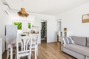 Eine moderne Küche mit weißen Schränken, einem Esstisch mit Stühlen, einem grauen Sofa und einer hängenden gewebten Leuchte.