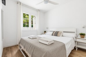 Une chambre à coucher comprenant un lit, une table de chevet, un ventilateur de plafond, une fenêtre avec des rideaux et une vue sur la verdure à l’extérieur.