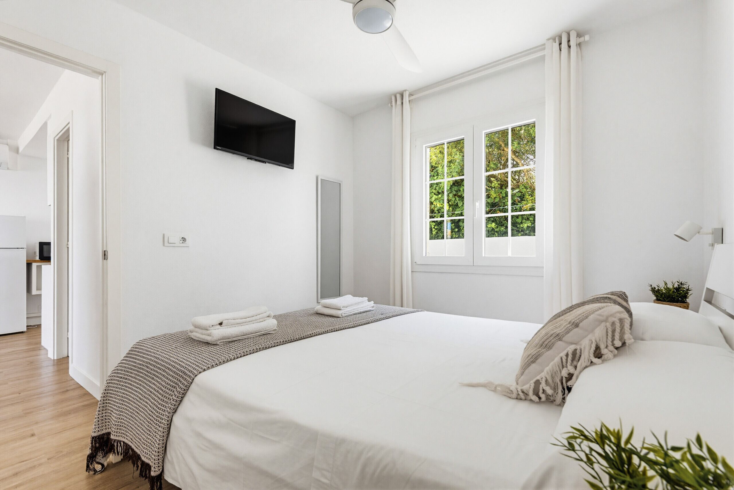 A bedroom with a bed, a TV mounted on the wall, a window with curtains, and a door leading to another room.