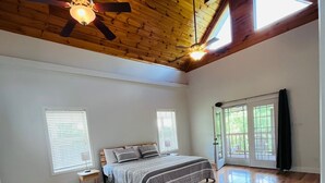 A spacious bedroom with a wooden floor, a bed with patterned bedding, a ceiling fan, and large windows allowing natural light. - Wears Valley Farmhouse with Wonderful Views, hot tub and firepit (Sevierville)
