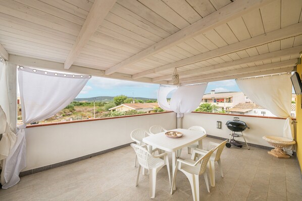 Un balcón techado con mesa y sillas, una parrilla y vistas a casas.