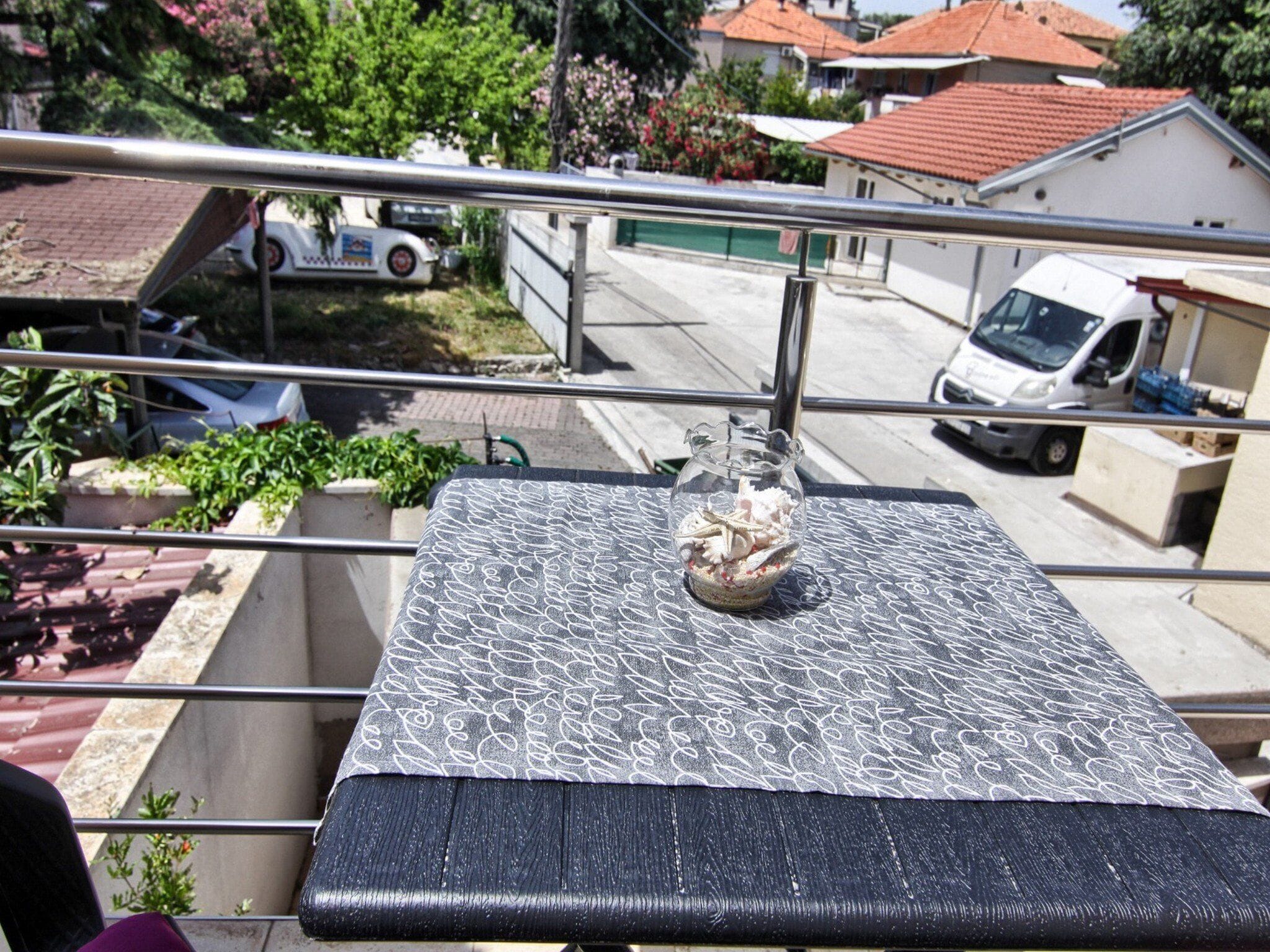 A balcony with a patterned tablecloth, a glass vase with white flowers, and a view of the street below.