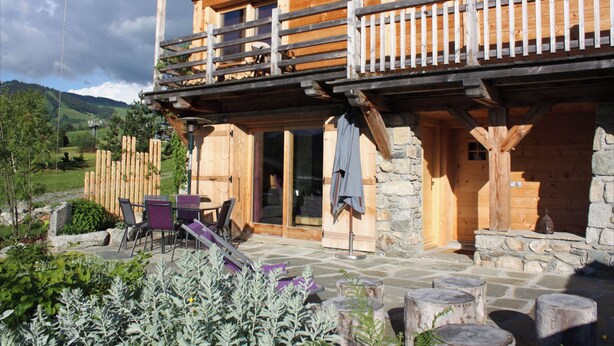 A wooden cabin with a balcony, stone walls, and a patio with outdoor furniture.