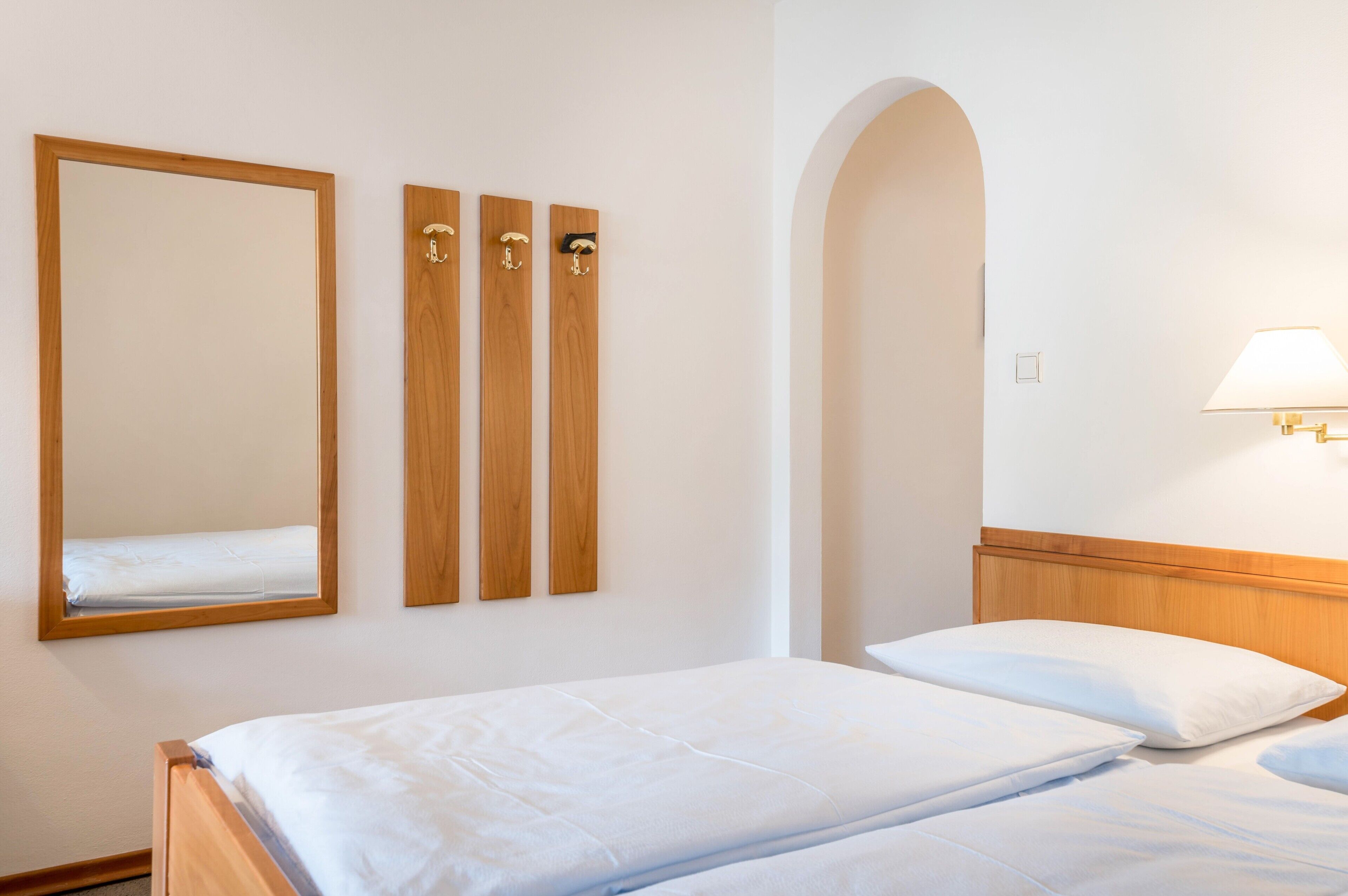 A hotel room with a bed, a mirror, and three wooden wall-mounted hooks.