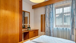 Un dormitorio con un armario de madera, una mesita de noche con espejo, una cama con ropa de cama blanca y una ventana con cortinas de encaje.