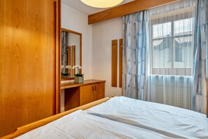 A bedroom with a wooden wardrobe, a bedside table with a mirror, a bed with white bedding, and a window with sheer curtains.