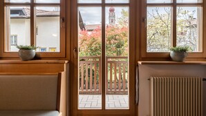 Quarto com porta de madeira, duas janelas com plantas em vasos, radiador e vista para um edifício com torre.