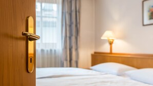 Quarto de hotel com porta de madeira, cama com roupa de cama branca, mesa de cabeceira com abajur e janela com cortinas.