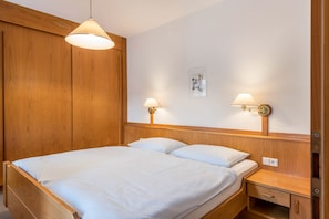 A wooden bed with white linens, two bedside lamps, and a small bedside table with a drawer.