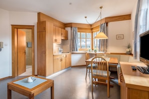 A compact kitchen with wooden cabinets, a dining area with a table and chairs, a TV, and a window with curtains.