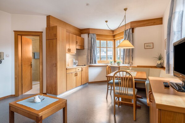 Une cuisine compacte avec des meubles en bois, un coin repas comprenant une table et des chaises, un téléviseur et une fenêtre avec des rideaux.