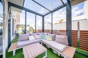 Un espace de vie extĂ©rieur moderne, dotĂ© dâune structure en verre, dâun canapĂ© modulable et dâune table en bois.