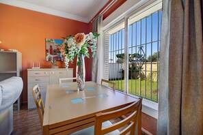 A dining area with a table, chairs, a vase with flowers, a painting on the wall, and a view through a window.