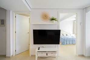 Une pièce avec un téléviseur posé sur un meuble, un lit recouvert d’une couverture à carreaux bleus et blancs, une plante en pot sur une étagère et une horloge murale.