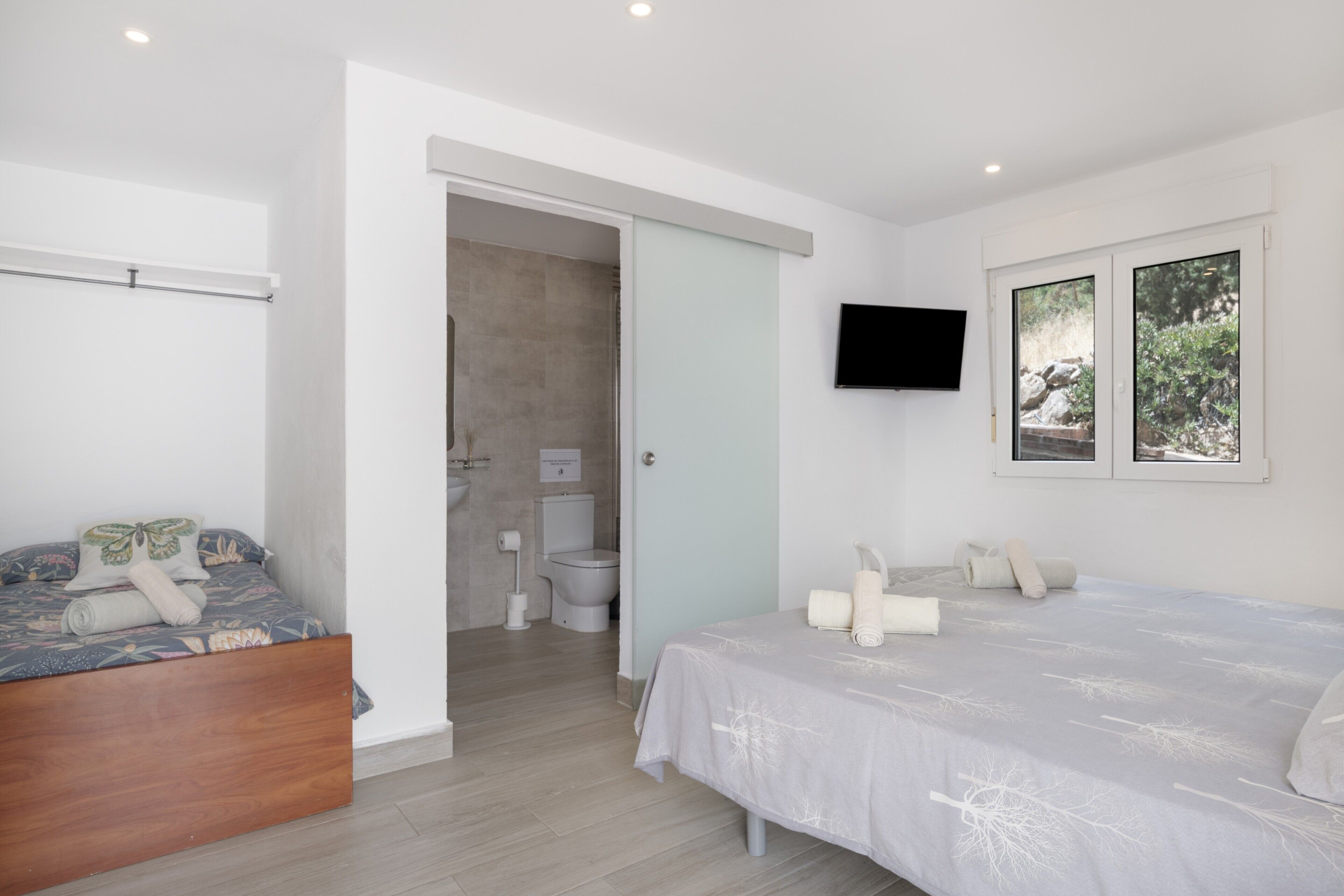 Un dormitorio con cama, televisor montado en la pared y ventana con vista a follaje.