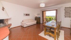 Ein Wohnzimmer mit Kamin, einer Couch, einem Esstisch mit Stühlen und Blick ins Freie.