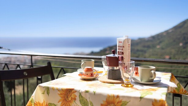Ein Tisch mit einer blumigen Tischdecke, einer Kaffeemaschine und Tassen auf einem Balkon mit Blick auf eine schöne Landschaft.