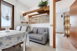 A living room with a grey sofa, a dining table with a blue tablecloth, a stone wall, and a wooden ceiling beam.