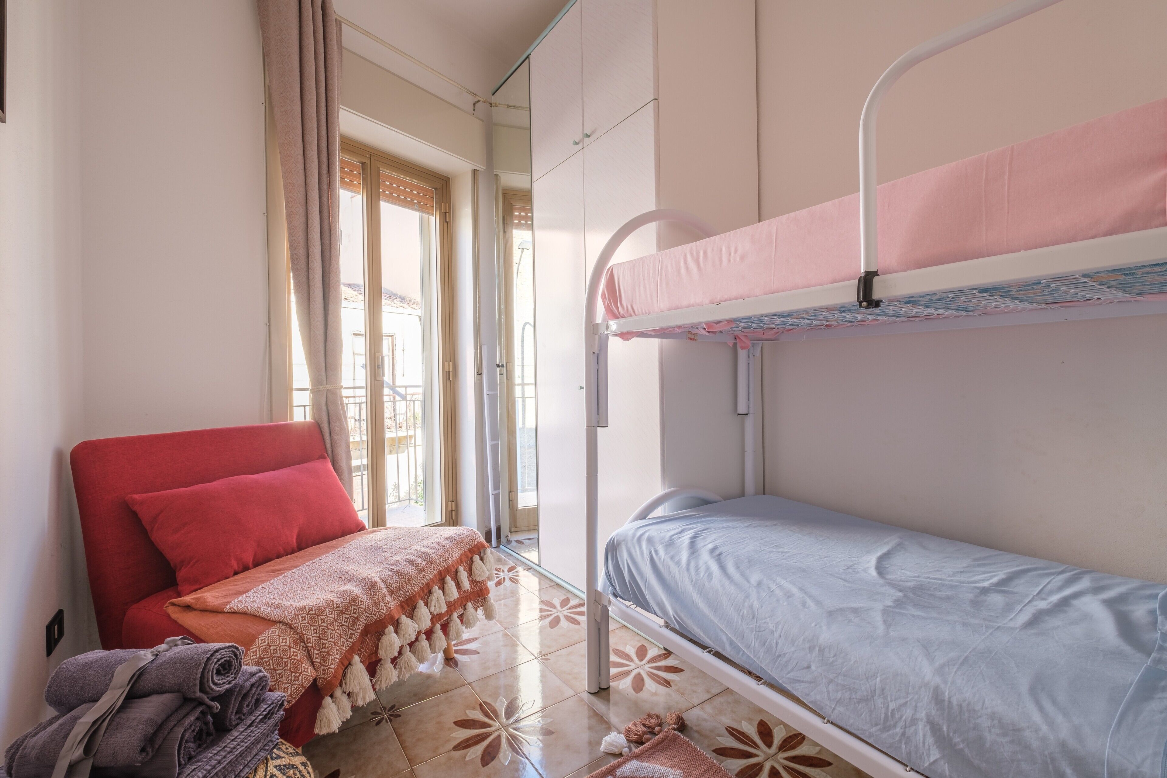Una habitación con una cama nido, una silla roja, un suelo estampado y una ventana con cortinas.