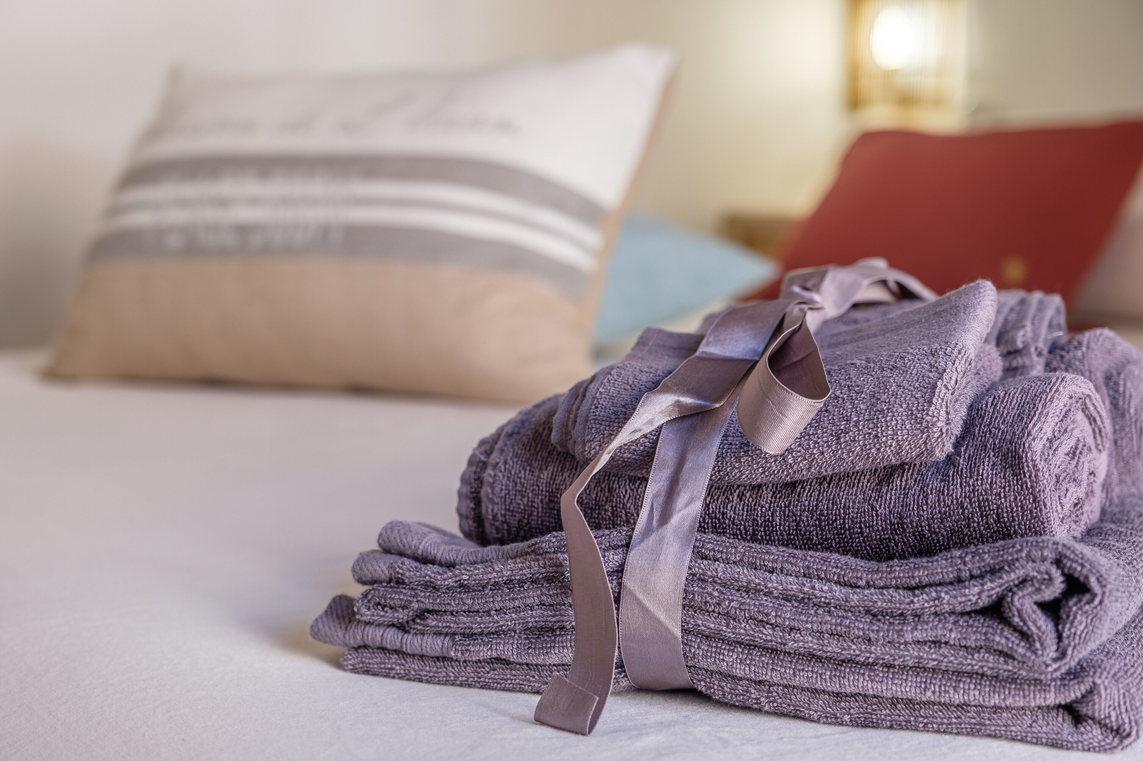 Ein ordentlich gefaltetes Handtuch mit Schleife auf einem Bett, ein Kopfkissen und ein rotes Buch im Hintergrund.