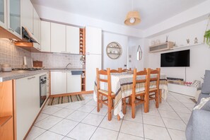 Cocina compacta con armarios blancos, una mesa de comedor con sillas, un televisor y una lámpara colgante.