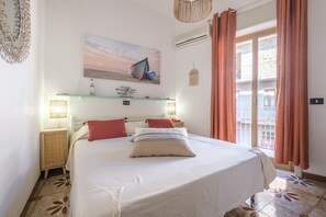Un dormitorio con cama, estante en la pared, ventana con cortinas y un cuadro de barcos.