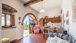 Une pièce confortable avec un lit, un bureau et une chaise, un coin cuisine doté de meubles en bois, et une vue sur l’extérieur à travers une porte en arc de cercle.