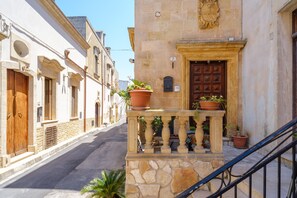 Eine enge Gasse mit steinernen Häusern, einer Holztür und einem Balkon mit Blumentöpfen.