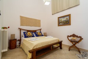A bedroom with a wooden bed, a bedside table, a radiator, a wall-mounted tapestry, and a framed picture.