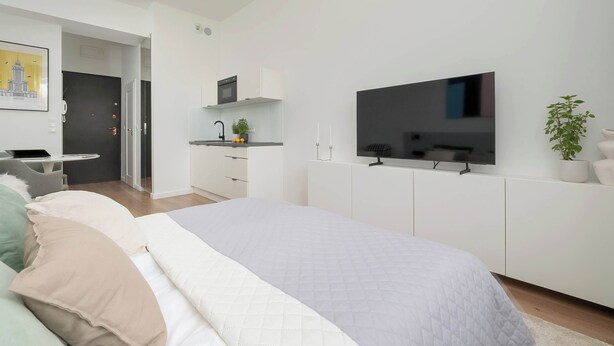 Een moderne slaapkamer met een bed, een televisie, een kitchenette en een deur naar een andere kamer.