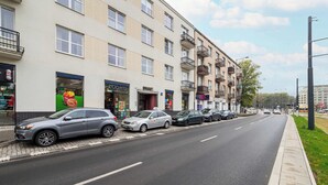 Een rij geparkeerde auto's langs een straat, met een gebouw dat meerdere balkons en een zichtbare winkelgevel heeft.