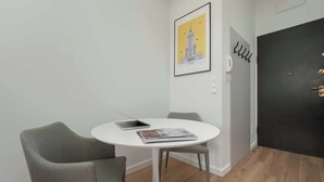 Een kleine kamer met een witte tafel, twee grijze stoelen, een lijst met een afbeelding van een gebouw, een zwarte deur en een kapstok met hangers.