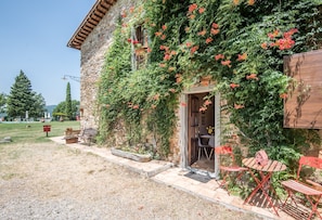 Un bâtiment en pierre aux murs recouverts de lierre, une porte en bois et un petit patio aménagé avec du mobilier d’extérieur.